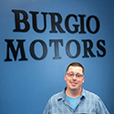Photo of Ray Burgio, owner of Lex-Burgio Motors against a blue wall with the words, Burgio Motors in black. 